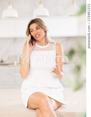 Irritated blonde talking on phone while sitting on kitchen table 118462252