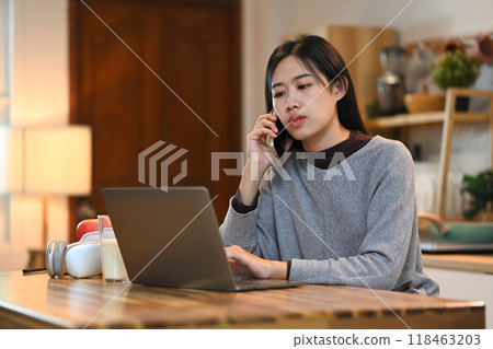 Young pretty woman using a laptop and talking on mobile phone in the kitchen 118463203