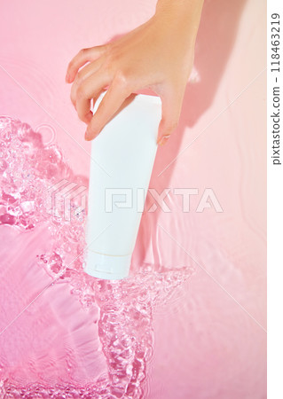 A woman's hand gently dips a white tube of skincare product into a pink water surface, creating a splash of bubbles and ripples. The image suitable for promoting skincare products or sunscreen. A woman's hand gently dips a white tube of skincare product into a pink water surface, creating a splash of bubbles and ripples. The image suitable for promoting skincare products or sunscreen. 118463219