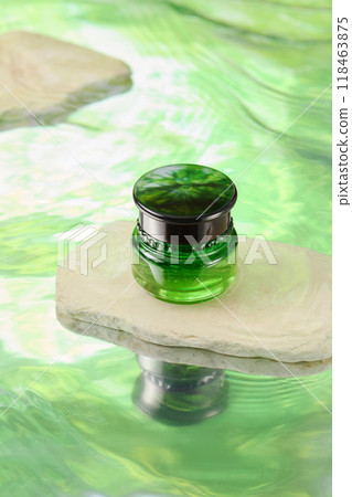 A sleek green unlabeled glass jar of cream sits on a white stone, reflecting off a rippling water green surface. Minimalist natural setting highlights the naturalness of the product 118463875