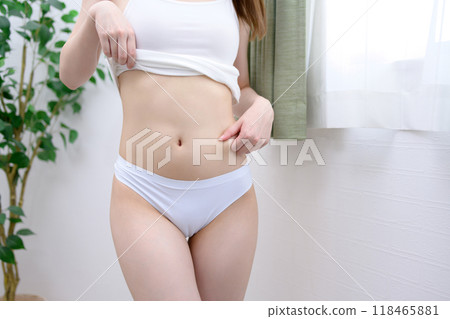 A young, slender woman with beautiful skin in underwear, pinching her thin belly by the window, daytime image 118465881