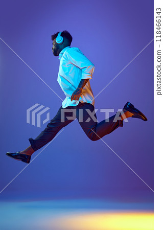 Young, energetic man in white shirt, black pants, and white headphones leaps mid-air in vibrant neon light against gradient background. Young, energetic man in white shirt, black pants, and white headphones leaps mid-air in vibrant neon light against gradient background. 118466143