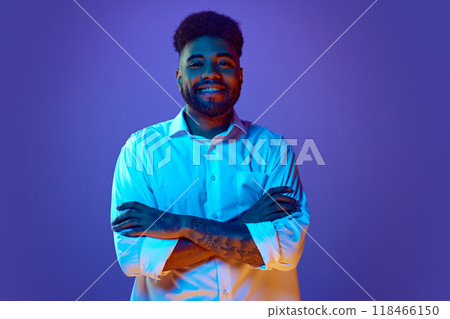 Crossing his tattooed arms, man in white shirt smiles warmly, looking at camera, in vibrant neon light against gradient background. 118466150