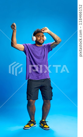 Joyful dancing African-American man in purple shirt wears white headphones against blue background. Listening music. Joyful dancing African-American man in purple shirt wears white headphones against blue background. Listening music. 118466152