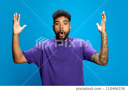African-American man, wearing purple shirt, looks surprised with his mouth open, raising his hands in playful gesture against blue background. 118466156