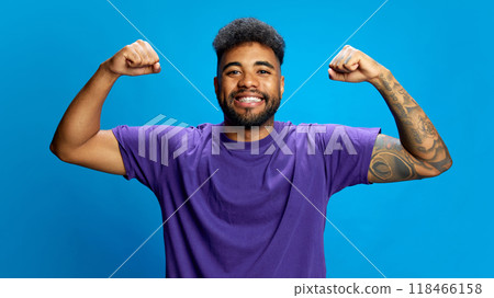 Young man in purple shirt smiling warmly. His arms raised, showing off his strength with flexing pose against vibrant blue background. 118466158