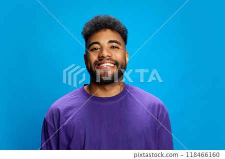 Young, bearded African-American man in stylish, vibrant purple shirt smiling warmly and looking at camera against blue background. 118466160