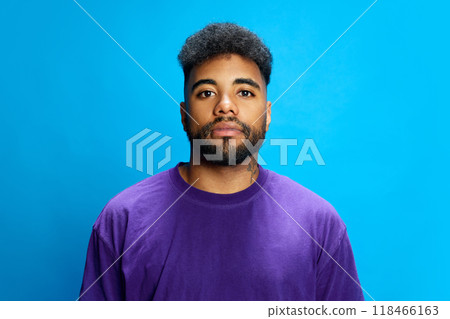 Bearded African-American man in purple shirt stands in relaxed pose, looking directly at camera against blue background. 118466163