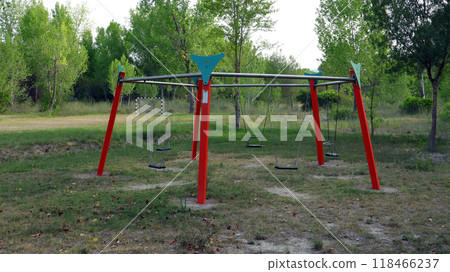 An empty playground features a large swing set with multiple swings, set in a peaceful park. Lush green trees surround the area, creating a calm atmosphere. An empty playground features a large swing set with multiple swings, set in a peaceful park. Lush green trees surround the area, creating a calm atmosphere. 118466237