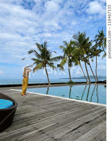 Middle aged blonde woman in yellow sarong and swimsuit standing by pool 118467135
