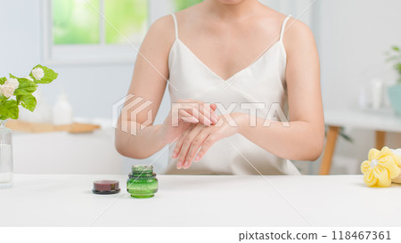 By the white table in bathroom scene is a woman with white dress spreading moisturizer on the back of her hand. A green cosmetic pot without label placed on white table, displaying and copy space By the white table in bathroom scene is a woman with white dress spreading moisturizer on the back of her hand. A green cosmetic pot without label placed on white table, displaying and copy space 118467361