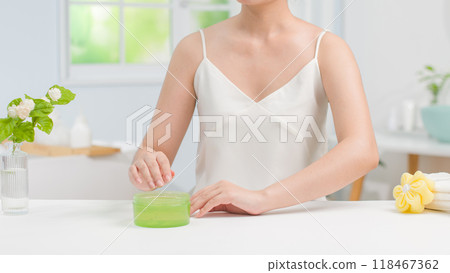 Woman in white dress sitting next to white table and preparing to apply lotion after shower. Mockup for advertising with a green unbranded cosmetic jar placed on table, space for product presentation Woman in white dress sitting next to white table and preparing to apply lotion after shower. Mockup for advertising with a green unbranded cosmetic jar placed on table, space for product presentation 118467362