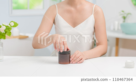 A woman with white strap dress sits in a bathroom after taking shower, unscrewing an unlabeled amber pot placed on a white table. Blank space for adding text and showing body care product 118467364
