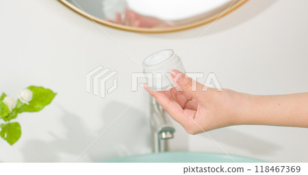 Close up shot at scene of an unlabeled white cream jar being held in female hand, over bathroom background. Cosmetic advertising photo was taken from front view, copy space Close up shot at scene of an unlabeled white cream jar being held in female hand, over bathroom background. Cosmetic advertising photo was taken from front view, copy space 118467369