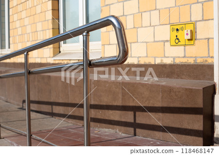 Metal railing is next to yellow sign that says handicap accessible with help button. Ramp for people in wheelchairs at the entrance to building with handrails for assistance Metal railing is next to yellow sign that says handicap accessible with help button. Ramp for people in wheelchairs at the entrance to building with handrails for assistance 118468147