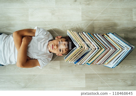 A cheerful boy lies on the floor, and above his head there is a large stack of books in the form of a roof. A cheerful boy lies on the floor, and above his head there is a large stack of books in the form of a roof. 118468645
