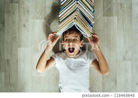 A cheerful boy lies on the floor, and above his head there is a large stack of books in the form of a roof. A cheerful boy lies on the floor, and above his head there is a large stack of books in the form of a roof. 118468648