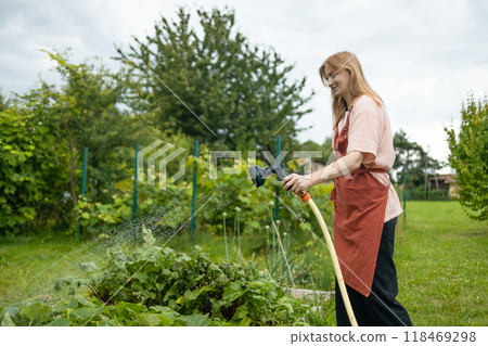 Happy woman watering fresh plants growing at home vegetable garden. Gardener taking care of plants at the backyard of her house. Concept of sustainability and growing organic 118469298