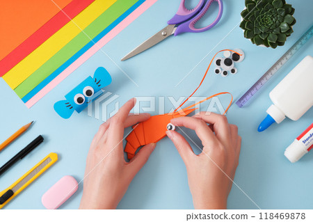 Making an origami shrimp out of orange paper. Women's hands attach an eye to a paper toy. Hobby idea. Making an origami shrimp out of orange paper. Women's hands attach an eye to a paper toy. Hobby idea. 118469878