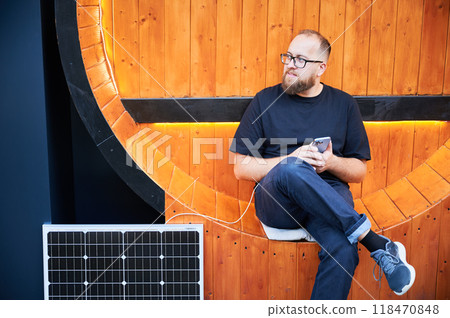 Bearded man using smartphone connected to photovoltaic solar panel. Integration of sustainable renewable energy into everyday life, demonstrating practical use of solar power for charging devices. 118470848