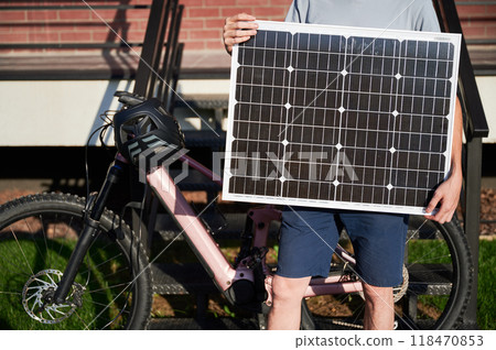 Close up view of person holding solar panel for charging, stands in front of brick building. Next to him pink electric mountain bike with black helmet resting on handlebars. Cropped view. Close up view of person holding solar panel for charging, stands in front of brick building. Next to him pink electric mountain bike with black helmet resting on handlebars. Cropped view. 118470853
