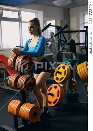 Young woman in activewear resting beside weights in a gym during daytime 118472249