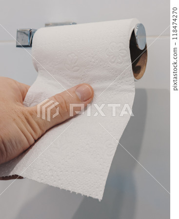 A hand pulls a sheet of toilet paper from a roll mounted on the wall in a bathroom 118474270