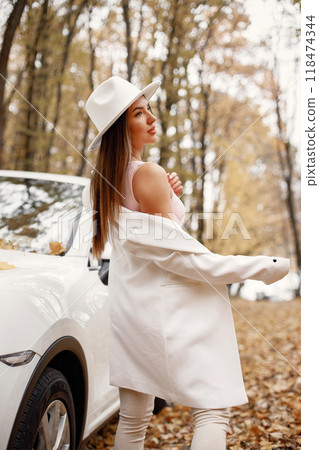 Portrait of woman in autumn forest, standing next to her white car. Brunette woman posing for a photo. Woman wearing white hat, pink t-shirt and white jacket. 118474344