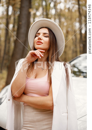 Portrait of woman in autumn forest, standing in front of her white car. Brunette woman posing for a photo. Woman wearing white hat, pink t-shirt. Portrait of woman in autumn forest, standing in front of her white car. Brunette woman posing for a photo. Woman wearing white hat, pink t-shirt. 118474346