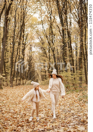 Young woman with little girl walking in autumn forest. Brunette woman play with her daughter. Girl wearing beige sweater and mother wearing white clothes. Young woman with little girl walking in autumn forest. Brunette woman play with her daughter. Girl wearing beige sweater and mother wearing white clothes. 118474350