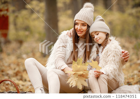 Young woman with little girl sitting on a fallen tree trunk in autumn forest. Brunette woman play with her daughter. Girl wearing beige sweater and mother wearing white clothes. Young woman with little girl sitting on a fallen tree trunk in autumn forest. Brunette woman play with her daughter. Girl wearing beige sweater and mother wearing white clothes. 118474351