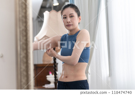 A woman wearing sportswear worries about her upper arms in front of a mirror A woman wearing sportswear worries about her upper arms in front of a mirror 118474740