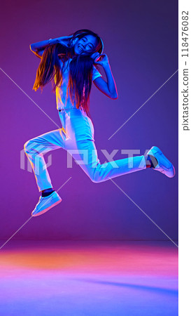 Girl, listening music in headphones, singing and leaping in motion of joy in neon light against gradient studio background. 118476082