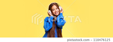Banner. Charming, creative young girl dressed warm sweater adjusting headphones against yellow background with negative space. Banner. Charming, creative young girl dressed warm sweater adjusting headphones against yellow background with negative space. 118476125