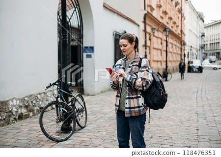Happy cheerful young woman with backpack walking on city street checks her smartphone. Portrait of beautiful 30s girl using smartphone outdoors. Urban lifestyle concept. Happy cheerful young woman with backpack walking on city street checks her smartphone. Portrait of beautiful 30s girl using smartphone outdoors. Urban lifestyle concept. 118476351