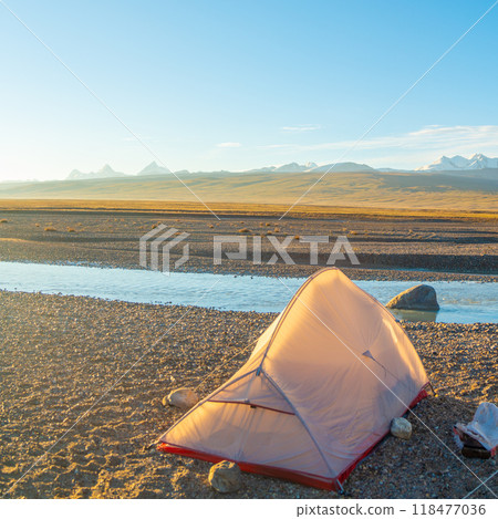 Tent stands on rocky ground next to a calm river, against a backdrop of majestic mountains and soft hills. The landscape combines the harshness of mountain nature and the tranquility of a quiet river 118477036