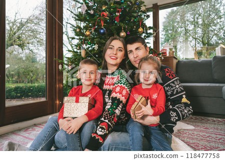 Smiling family portrait in festive sweaters sitting with gifts under Christmas tree at home during holiday. Happy parents and kids cuddling, feeling excited. Family time together. Happy childhood. Smiling family portrait in festive sweaters sitting with gifts under Christmas tree at home during holiday. Happy parents and kids cuddling, feeling excited. Family time together. Happy childhood. 118477758