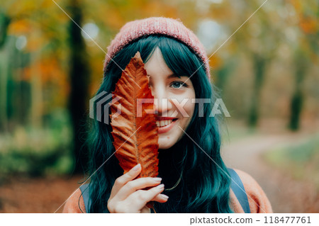 Portrait of Young smiling woman in hat partially covering her face with striking red fallen leaf enjoying autumn walk in forest. Feeling harmony, reunion with nature. Relaxing, personal fulfilment 118477761