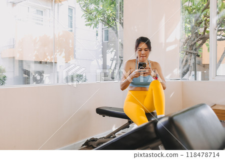 Lazy woman in Yellow Blue fitness outfit sitting and using mobile in the gym. 118478714