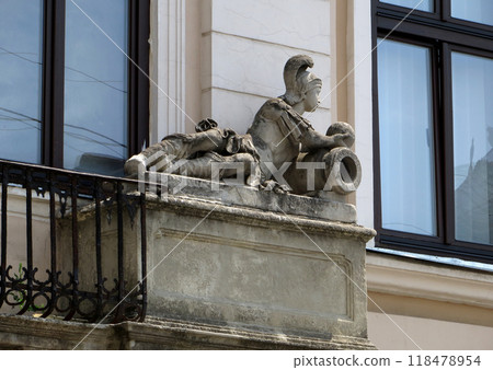 Reclining figure of a Roman soldier with cannon and a cannonball. Stone statue on the old building. 118478954