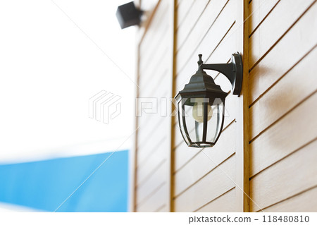 A stylish outdoor lantern mounted on wooden wall, providing warm glow and enhancing ambiance of space. This elegant fixture complements modern architecture beautifully A stylish outdoor lantern mounted on wooden wall, providing warm glow and enhancing ambiance of space. This elegant fixture complements modern architecture beautifully 118480810