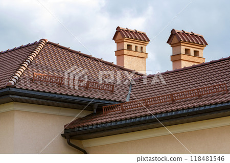Top of new big spacious modern house with shingled red roof, two high chimneys, plastered walls and snow protection fence against bright blue sky background. Professionally done construction concept. 118481546