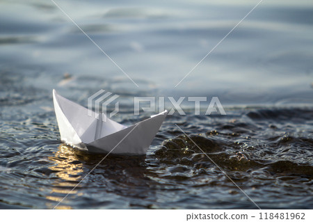 Close-up of simple small white origami paper boat floating in blue clear river or sea water under bright summer sky. Beauty of nature, freedom, dreams and fantasies concept. Close-up of simple small white origami paper boat floating in blue clear river or sea water under bright summer sky. Beauty of nature, freedom, dreams and fantasies concept. 118481962