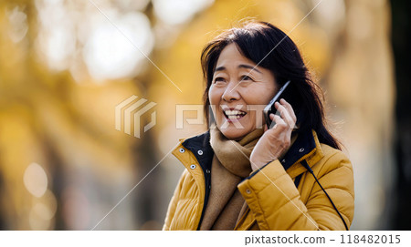 An asian woman in a yellow coat joyfully talks on her phone while walking through a park surrounded by autumn trees An asian woman in a yellow coat joyfully talks on her phone while walking through a park surrounded by autumn trees 118482015
