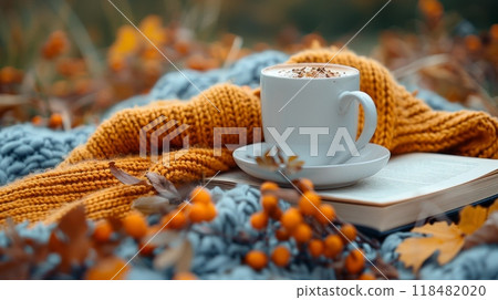 Autumn or fall season. Seasonal composition in the park in dry autumn foliage. A colorful knitted scarf, a cup of hot coffee in a sweater and an old book. Sea buckthorn twig, warm toned 118482020