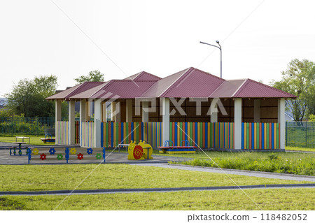 Beautiful playground in kindergarten with bright new alcove with red tile roof and multicolored low fence, green lawn, swings, slides and sandbox. Perfect place for children activities outdoors. 118482052