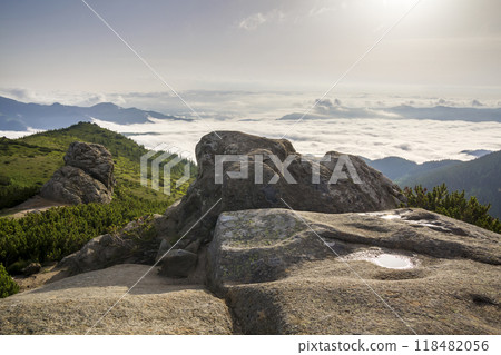 Beautiful summer mountain panorama at dawn. Huge boulder with rain water puddle on green rocky mountain top on background of foggy valley filled with white puffy clouds and clear blue copy space sky. 118482056