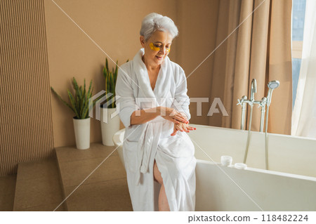 Mature adult old woman applying moisturizing body lotion on hands. Senior lady rubbing nourishing anti age cream doing morning daily skincare routine in bathroom. Beauty treatment spa body skin care Mature adult old woman applying moisturizing body lotion on hands. Senior lady rubbing nourishing anti age cream doing morning daily skincare routine in bathroom. Beauty treatment spa body skin care 118482224