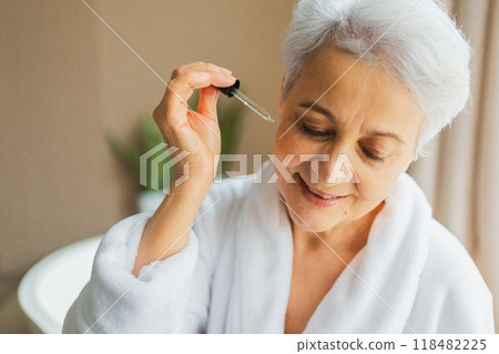 Woman applying natural drops cosmetics oil to face skin. Mature adult old woman applying nourishing anti age serum with pipette doing daily beauty routine. Senior woman gets kin scare facial treatment Woman applying natural drops cosmetics oil to face skin. Mature adult old woman applying nourishing anti age serum with pipette doing daily beauty routine. Senior woman gets kin scare facial treatment 118482225