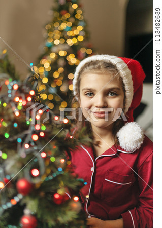 Happy girl of 7 years old in red pyjamas and Santa Claus hat next to Christmas tree with side of garlands with bokeh Happy girl of 7 years old in red pyjamas and Santa Claus hat next to Christmas tree with side of garlands with bokeh 118482689